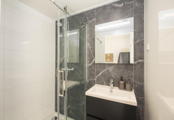 Baño moderno en una posada con ducha de vidrio, lavabo oscuro, gran espejo y azulejos efecto mármol.