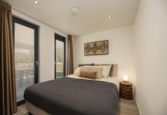 Modernes Schlafzimmer in einer Lodge mit Doppelbett, großen Fenstern, braunen Vorhängen und warmem Licht.