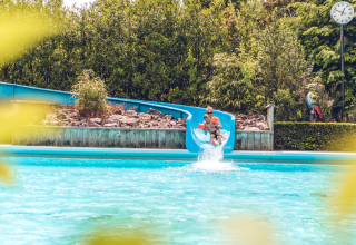 Un adulto y un niño bajan por un tobogán azul hacia la piscina en un alojamiento glamping bajo el sol.
