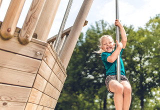 Una niña sonriente juega en un tubo resbaladizo en el parque infantil de una cabaña, rodeada de árboles.