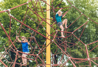 Dos niños juegan y trepan una gran estructura de cuerdas afuera en una lodge, rodeados de árboles verdes.