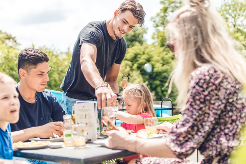 Familie genießt eine Mahlzeit im Freien beim Glamping, umgeben von Natur und sonnigem Wetter.