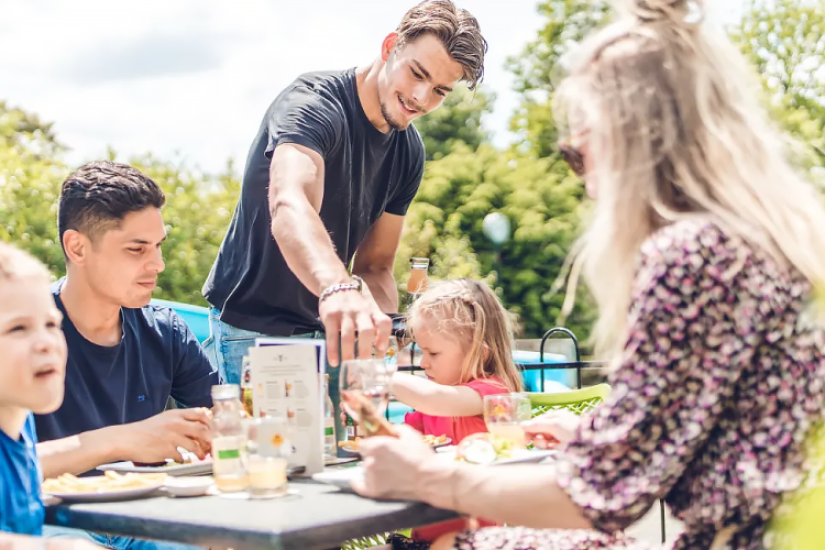 Family enjoys an outdoor meal at a glamping site, smiling together in sunny weather and nature.