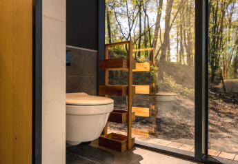 Modern bathroom with toilet and wooden shelf, glass wall offering forest view at ÖÖD Suite in Germany.