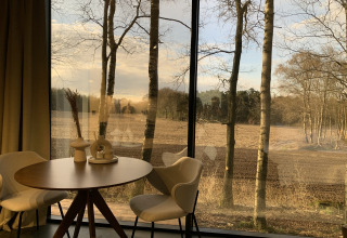 Modern interieur met ronde tafel en stoelen naast grote ramen, uitkijkend op een veld en bomen.