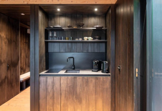 Modern kitchenette in Nokken Forest Cabin at Wilsumer Berge, Germany, featuring dark wood and open shelves.