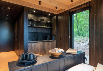 Cabane moderne en bois sombre avec petite cuisine et grande fenêtre offrant vue sur la forêt verdoyante.