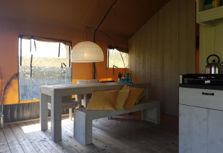 Interior de una tienda safari con mesa y bancos de madera, cojines amarillos y luz natural abundante.