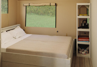 Vista interior de tienda safari en Veluwepark de Bosgraaf, Países Bajos, con cama y estantería blanca.