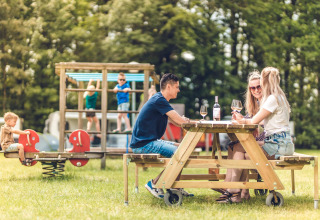 Tres adultos disfrutan vino en una mesa de picnic mientras los niños juegan en un parque en la naturaleza.