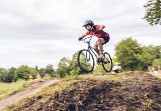 Un niño con casco salta en bicicleta sobre una loma de tierra en un alojamiento glamping con árboles detrás.