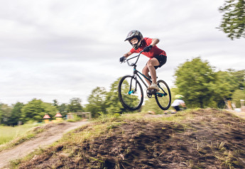 Een kind met helm maakt een sprong op de fiets bij een glampingverblijf met bomen en speelplek achteraan.