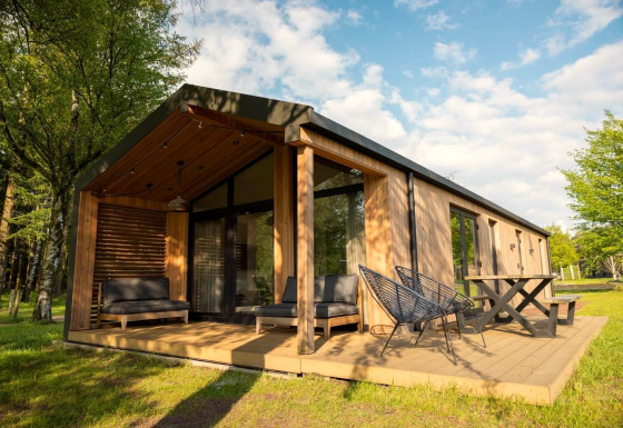 Lodge moderne en bois avec grande terrasse, mobilier extérieur et environnement verdoyant sous un beau ciel.