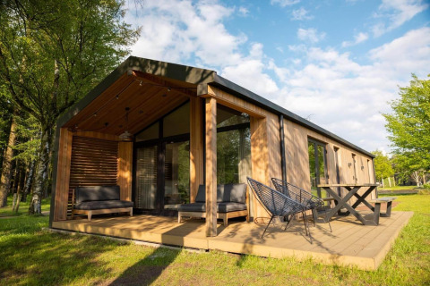 Moderne houten lodge met groot terras, buitentoelen en veel groen gras tussen de omliggende bomen.