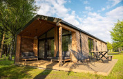 Lodge moderno de madera con terraza grande, sillas exteriores y rodeado de césped verde y árboles altos.