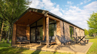 Lodge moderno de madera con terraza grande, sillas exteriores y rodeado de césped verde y árboles altos.