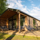 Modern wooden lodge with spacious deck, outdoor seating, lush green grass, and trees in sunlight.