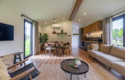 Salon moderne en lodge avec grandes fenêtres, sol en bois, mobilier confortable et cuisine ouverte.