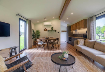 Salon moderne en lodge avec grandes fenêtres, sol en bois, mobilier confortable et cuisine ouverte.