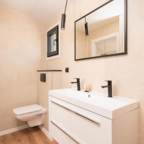 Moderne lodgebadkamer met zwevend toilet, dubbele wastafel, zwarte kranen en grote spiegel aan de muur.