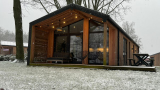 Modern wooden lodge at Wilsumer Berge, Germany, with a snowy yard and bare winter trees in the background.