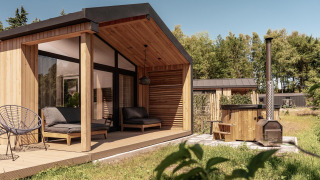 Modern wooden lodge with covered terrace, outdoor seating, and hot tub set amidst lush green surroundings.