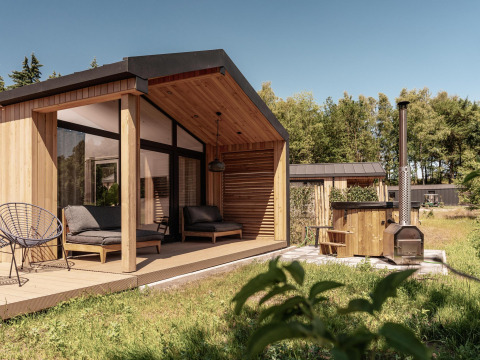 Moderne Holzlodge mit überdachter Terrasse und Outdoor-Badewanne, umgeben von grüner Natur und Bäumen.