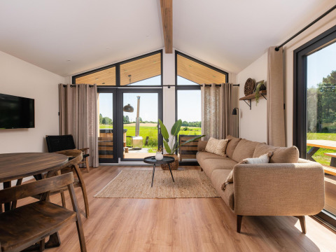 Salon de lodge moderne avec grandes fenêtres, canapé beige et vue sur des champs verts à l'extérieur.