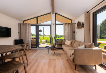 Soggiorno di un lodge moderno con grandi finestre, divano beige e vista su campi verdi all’esterno.