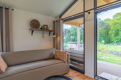 Knusse lodge met zetel, wandplank, grote ramen en zicht op houten terras en groene tuin buiten.