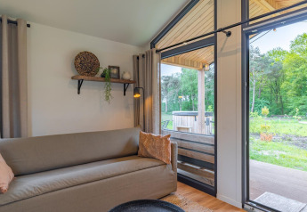 Intérieur de lodge avec canapé, étagère murale, grandes fenêtres et terrasse ouverte sur la nature verte.