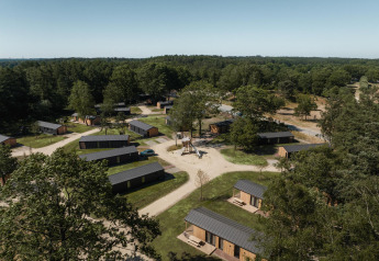 Luchtfoto van een lodge met kleine houten chalets, omringd door bos en wandelpaden onder een heldere hemel.