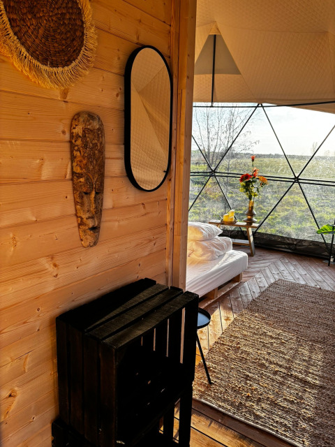 Intérieur de la maison nature Cast Away Dome, murs en bois, lit, tapis et vue sur la campagne néerlandaise.