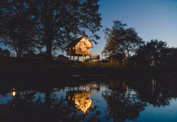 Beleuchtetes Baumhaus im Holiday Park Mölke, Niederlande, spiegelt sich abends im ruhigen Wasser.