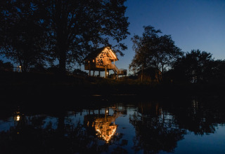 Casa del árbol iluminada en Holiday Park Mölke, Países Bajos, reflejada en el agua al anochecer.