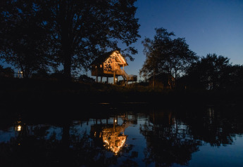 Oplyst træhus i Holiday Park Mölke, Holland, reflekteres i vand om aftenen med omgivelser af træer.