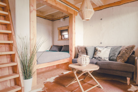 Cozy tree house living area with grey sofa, round table, bed nook, and wood accents in Holiday Park Mölke.