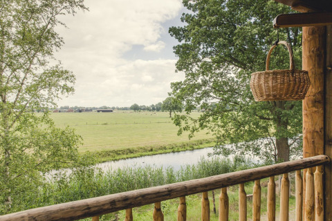 Uitzicht vanuit een boomhut in Holiday Park Mölke, Nederland, met weiland, rivier en mand op balkon.