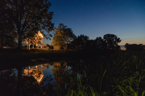Illuminated tree house by a pond at Holiday Park Mölke, Netherlands, surrounded by trees at dusk.