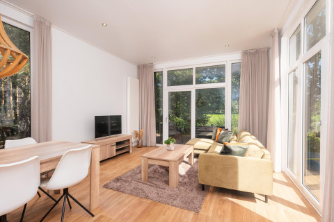 Lyst glampingophold med komfortabel stue, spisebord, sofa og store vinduer med udsigt til naturen.
