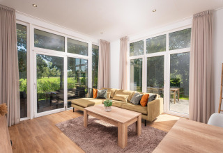 Licht glampingverblijf met grote ramen, moderne bank, houten tafel, gordijnen en uitzicht op het groen.