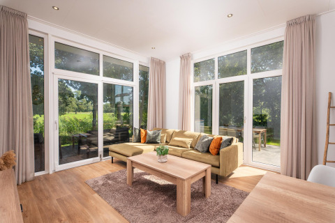 Licht glampingverblijf met grote ramen, moderne bank, houten tafel, gordijnen en uitzicht op het groen.