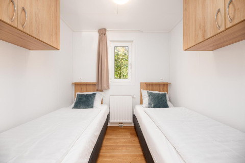 Lichte glampingkamer met twee aparte bedden, houten kasten, witte muren en een raam in het midden.
