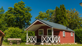 Red wooden cabin with porch at Scandinavian Lodge, surrounded by lush trees and a bright blue sky.