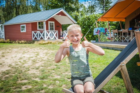 Junge sitzt vor einer skandinavischen Hütte im Holiday Park Mölke in den Niederlanden und zeigt Daumen hoch.