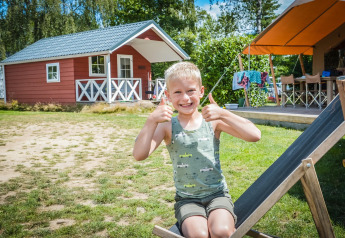Niño sentado afuera de una cabaña escandinava en Holiday Park Mölke, Países Bajos, mostrando pulgares arriba.
