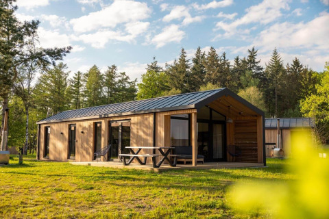 Cabaña de madera moderna con gran terraza y mesa de picnic en un entorno boscoso y soleado.