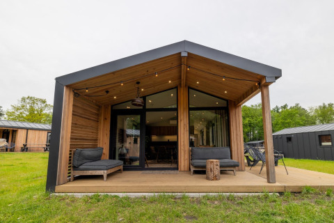 Moderne houten lodge met overdekt terras, zwarte loungezetels en grasveld in een recreatiepark.