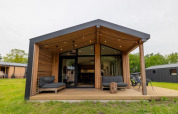 Moderne houten lodge met overdekt terras, zwarte loungezetels en grasveld in een recreatiepark.