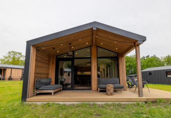 Moderne houten lodge met overdekt terras, zwarte loungezetels en grasveld in een recreatiepark.